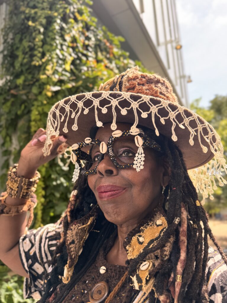 Portrait of Gina Renee wearing a wide-brimmed hat, shell-adorned glasses and intricately layered African-inspired clothing while posing outdoors beside a wall covered in green vines.