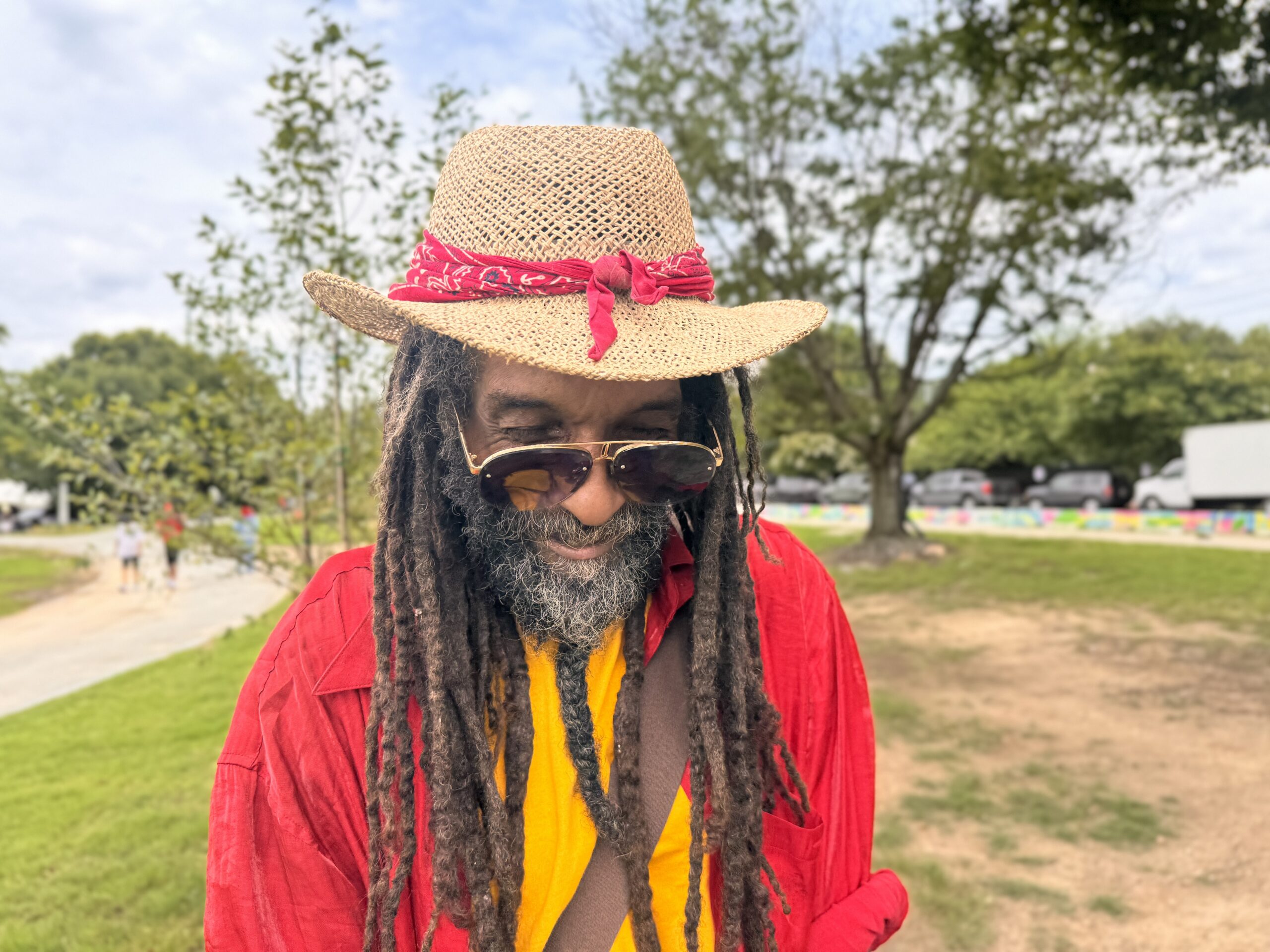 Portrait of musician Yesi standing outdoors in a park wearing a straw hat with a red bandana, sunglasses, a red shirt and long dreadlocks while looking down.